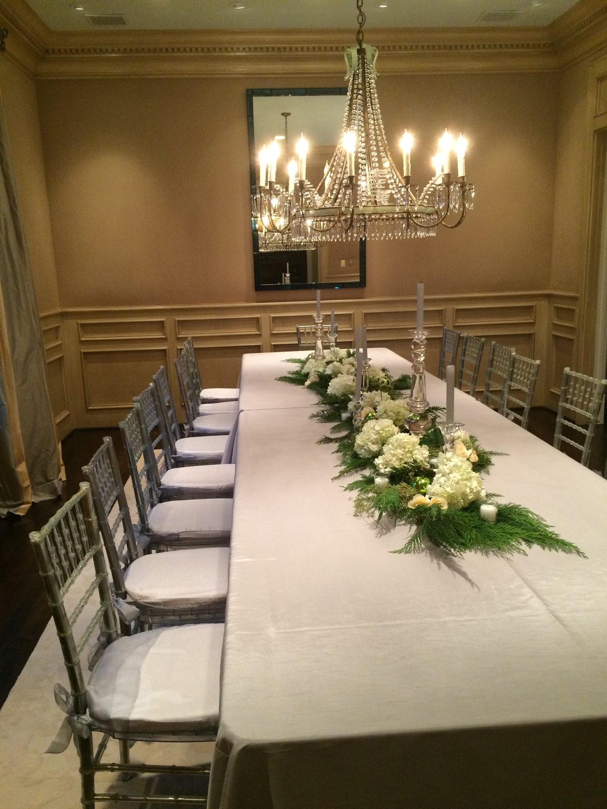 A long dining table set with a white floral and greenery runner under a crystal chandelier